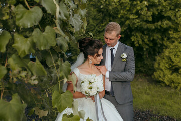 A bride and groom are standing in a lush green field, with the bride holding a bouquet of flowers....
