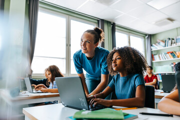 Female teacher bending near curly hair girl sitting with laptop near desk in classroom