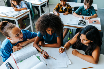 High angle view of teacher helping female pupils studying while sitting near desk in classroom