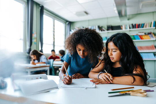 Curly hair girl writing while sitting with female classmate near desk in classroom at elementary school