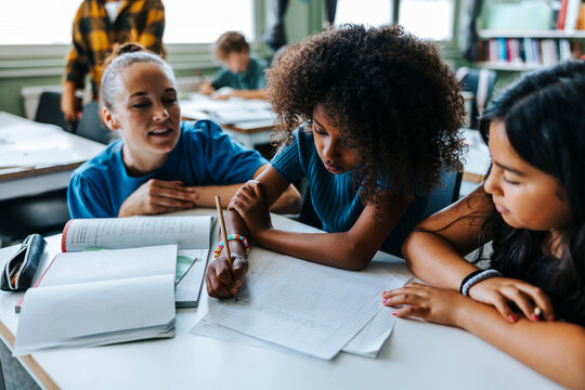Professor crouching near female student writing on paper while sitting beside classmate in classroom at elementary schoo