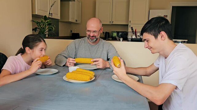 Happy family bite boiled corn holding cob in their hands home in kitchen. Delicious snack and good source of natural vitamins. Children and father eat corn and smile at each others faces