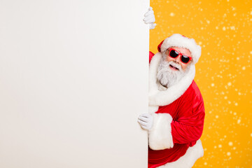 Portrait of funny funky christmas father in red hat headwear look behind white wall poster advertise winter x-mas shopping seasonal miracle sales discounts isolated over bright color background