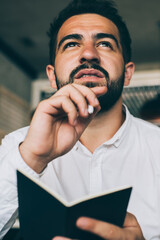 Contemplative male manager pondering on idea for business startup project during working process in coffee shop, pensive bearded man pondering on accounting information and strategy checklist