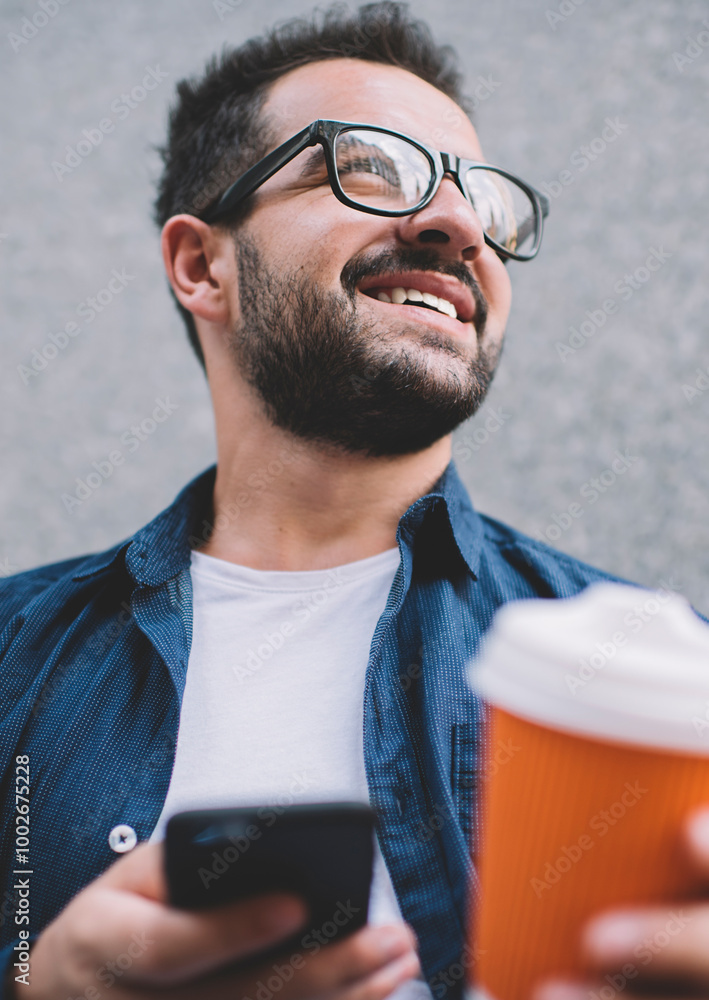 Poster below view of millennial bearded male blogger with takeaway cup in hand looking away and smiling dur