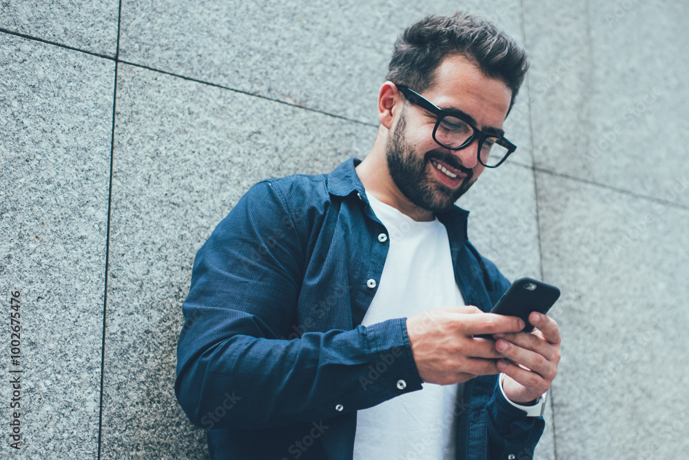 Poster positive male blogger in optical spectacles for vision correction standing near city wall and typing