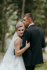 A bride and groom are posing for a picture in a forest. The bride is wearing a white dress and the groom is wearing a black suit. They are both smiling and hugging each other