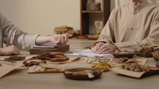 The patient pulse is taken by stretching and placing their palm on fabric-covered wooden board, while several herbs are on display in a traditional medicine clinic, as depicted in the template video.