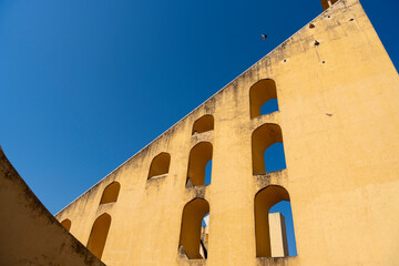  a section of Jaipur’s Jantar Mantar observatory, featuring a large yellow wall with arched openings and a curved staircase. The geometric design is highlighted against a blue sky.
