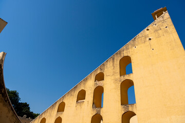  a section of Jaipur’s Jantar Mantar observatory, featuring a large yellow wall with arched openings and a curved staircase. The geometric design is highlighted against a blue sky.