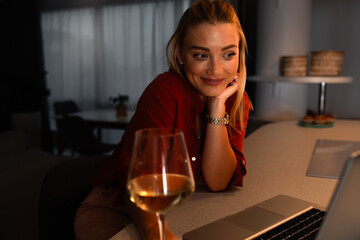 Smiling woman working at home using laptop while drinking wine at night.