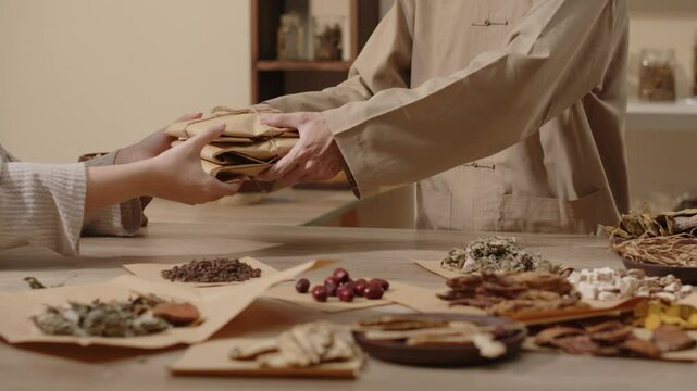A table full of herbs is placed in a traditional medicine room alongside a herbalist who is giving out packages of herbs wrapped in brown paper to a customer in traditional medicine video template.