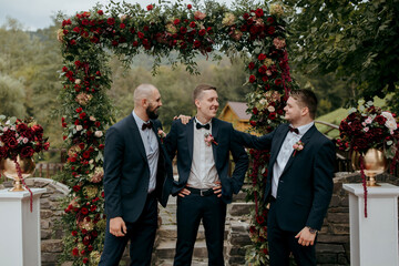 Three men in suits stand under a red archway, posing for a photo. The archway is decorated with flowers, and there are vases on the ground. Scene is celebratory and festive