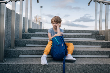 Little boy, schoolboy, with a backpack sits on stairs and is sad