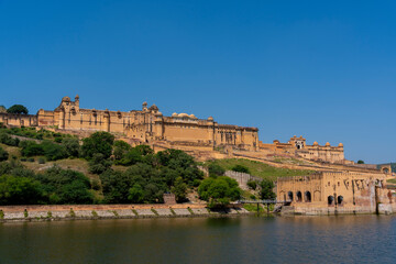 the majestic Amber Fort in Jaipur, Rajasthan, overlooking Maota Lake. The fort’s golden-yellow sandstone facade stands out against the lush greenery and clear blue sky.