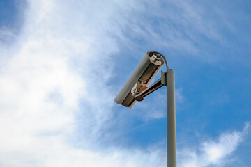 A camera mounted on a pole in the sky. The sky is blue with some clouds. The camera is pointed...