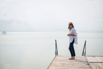 Fototapeta premium Full length of a blond haired woman standing on the pier by the lake