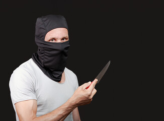 A male in a black mask and grey shirt grips a knife tightly while looking intently on dark...