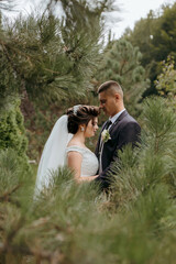 A bride and groom are standing in a forest, surrounded by trees. The bride is wearing a white dress and the groom is wearing a suit. They are embracing each other, looking into each other's eyes