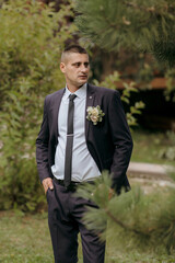A man in a suit and tie stands in a forest. He is wearing a flower in his lapel