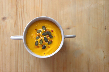 Bowl of creamy pumpkin and potato soup, with pumpkin seeds and chilli flakes toppings. Top view, wooden background.