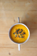 Bowl of creamy pumpkin and potato soup, with pumpkin seeds and chilli flakes toppings. Top view, wooden background.
