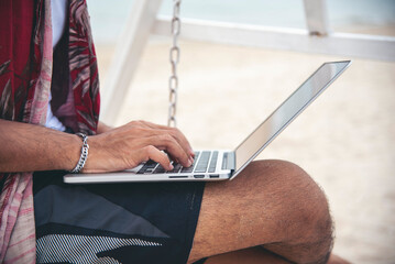 Man traveler use laptop on summer beach blue sky. Asian man wear casual short pants tropical shirt style relax on the beach in summertime. Man typing laptop computer  holiday business trip