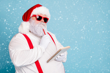 Close up photo of focused funny funky fat santa claus hold notebook think about wish list plan decide what choose look thoughtful wear stylish trendy suspenders isolated over blue color background