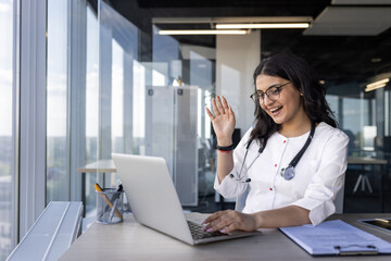 Female doctor engaging in virtual consultation, smiling waving at laptop. Wears stethoscope, suggesting medical context. Office space creates professional atmosphere. Concept of remote healthcare