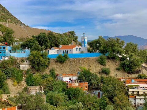 Tariq Ibn Ziyad Mosque perched on a hillside, features white and blue architecture surrounded by lush greenery and traditional Moroccan homes, set against a scenic mountainous backdrop.