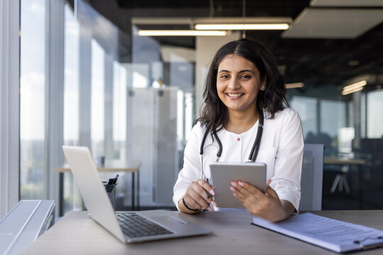 Young female doctor smiling at camera while holding tablet in modern office setting. Stethoscope around neck indicates professional healthcare role - Powered by Adobe