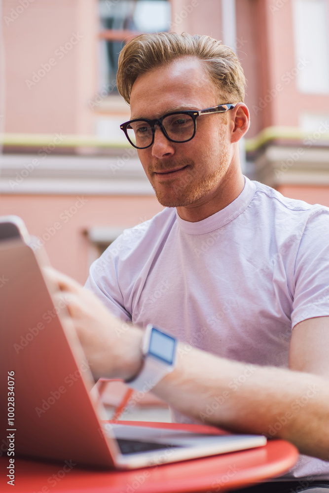 Poster positive man in classic eyewear sharing media files between laptop and smartphone devices using blue