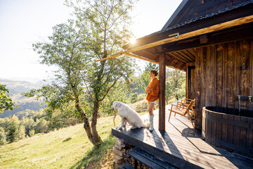 Woman in orange sweater with her dog on a cozy cabin porch. Sunlight filters through the trees,...