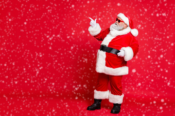 Full length profile side photo of charming santa claus in eyewear eyeglasses showing promo wearing bright costume belt isolated over red background