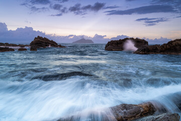 At dusk. Gentle waves lap against smooth, rounded rocks in a tranquil cove. The sky is painted in soft hues of pink and purple, reflecting on the calm water. Toucheng, Yilan, Taiwan.