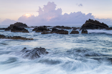 At dusk. Gentle waves lap against smooth, rounded rocks in a tranquil cove. The sky is painted in soft hues of pink and purple, reflecting on the calm water. Toucheng, Yilan, Taiwan.