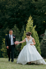 A bride and groom are walking together in a park. The bride is holding a bouquet and the groom is holding her hand. Scene is happy and romantic