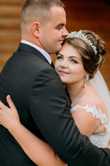 A bride and groom are hugging each other. The bride is wearing a tiara and the groom is wearing a...