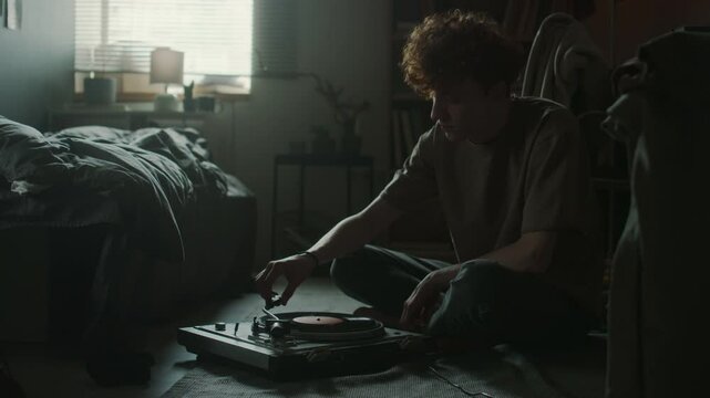 Melancholic young man putting on vinyl record while sitting on floor in front of player in his room