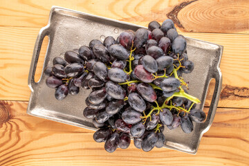 One bunch of ripe black grapes with metal tray on wooden table, macro, top view.
