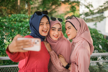 Three joyful women take a selfie in a lush outdoor setting, wearing colorful hijabs