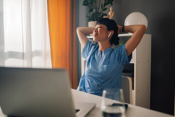 Relaxed asian woman nurse in blue scrubs stretching while sitting at desk