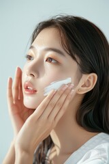 Asian model woman applies cream to face in beauty studio. Central composition on white background. Facial care routine shows skin moisturizing, pampering at home spa. Gentle hands apply lotion.