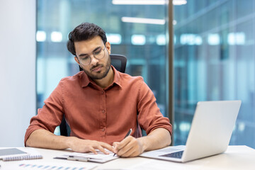 Young man in thoughtful expression working on important paperwork at desk. Displays focus and...