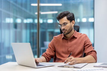 Confident man wearing glasses concentrating on laptop while taking notes. Engaged in serious paperwork, using digital tools for efficient work. Modern office environment enhances productivity focus.