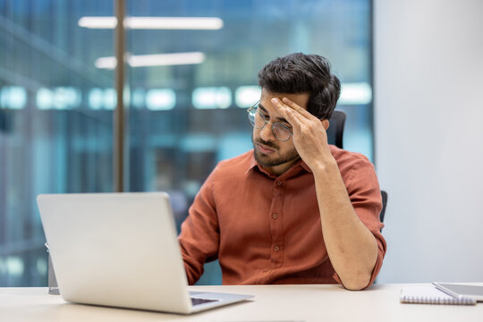 Young professional wearing glasses and brown shirt appears stressed while focusing on laptop in office. Man sits at desk with hand on forehead, indicating mental fatigue or work pressure.