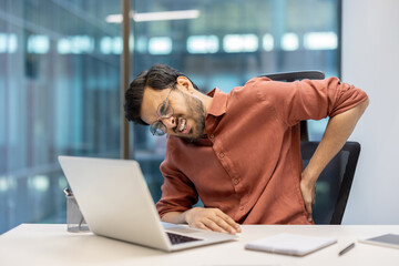 Man sitting at desk using laptop experiencing back pain. Wearing glasses shirt. Scene depicts discomfort from prolonged sitting. Represents office work, health issues, and need ergonomic solutions.