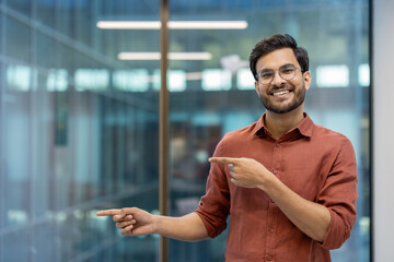 Smiling young man in glasses and casual attire pointing confidently in office setting. Represents positivity, confidence, and professional demeanor in modern workspace.