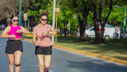 Two Latina women checking smartwatches while running outdoors