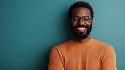 Fototapeta premium A cheerful man with a beard and glasses, dressed in an orange sweater, smiles warmly against a teal background, showcasing confidence and positivity.